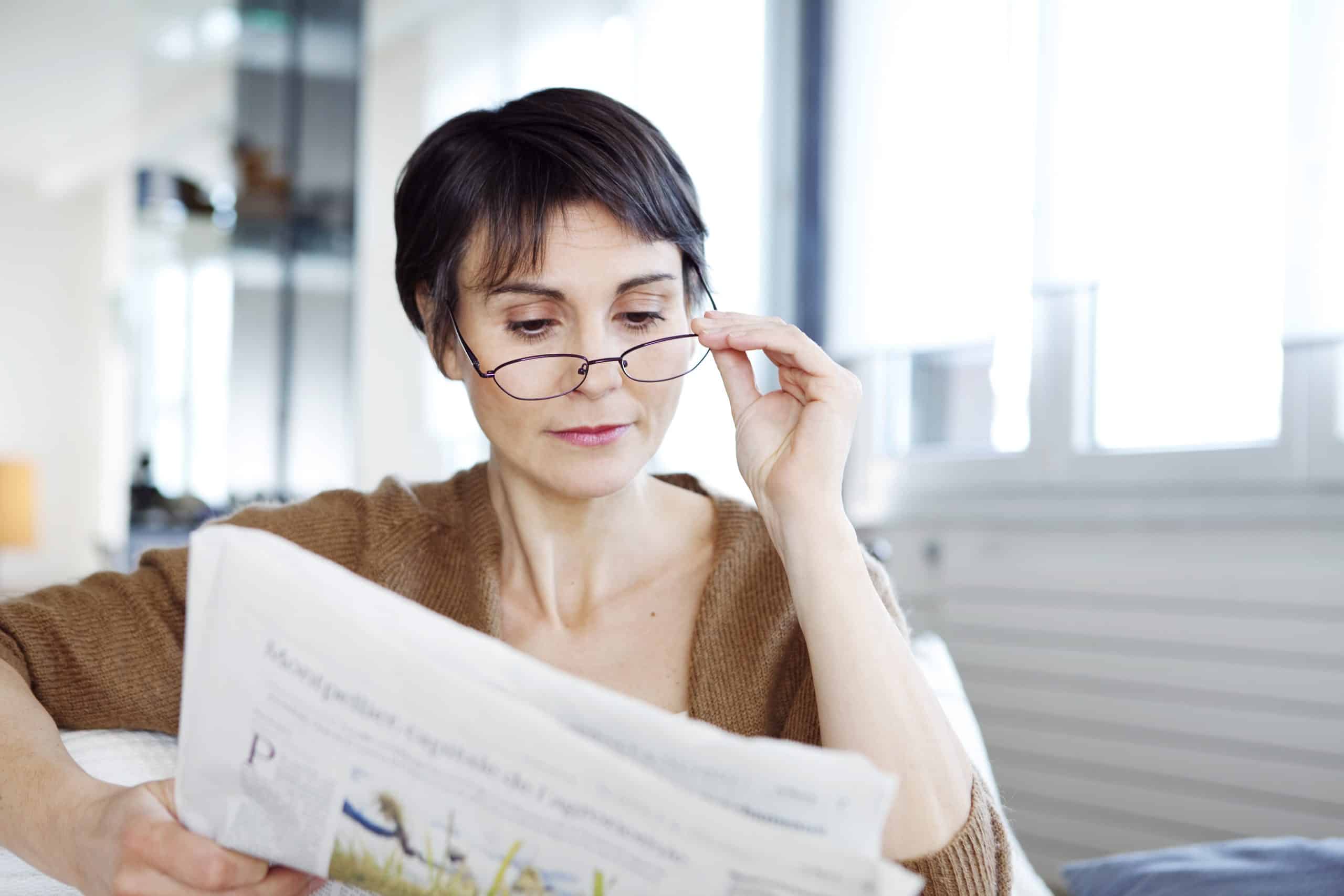 Woman reading newspaper pulling back glasses trying to decipher small text